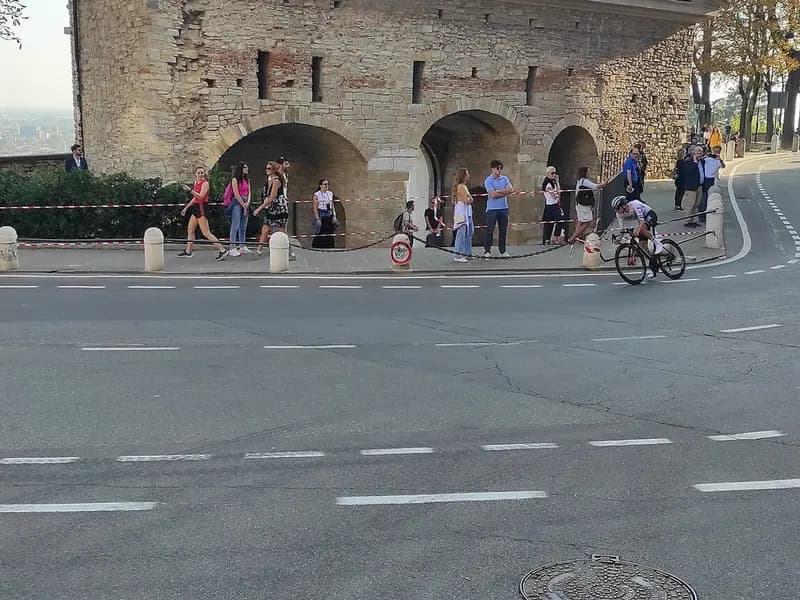 A lone cyclist rounding a bend beneath an old stone wall with spectators and a police motorcycle beside the road, photographed during Il Lombardia on 7 October 2023, a file photograph of the race and not of Paul Seixas or of Saturday's finish