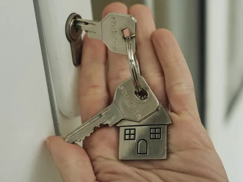A hand holding a set of house keys on a house shaped fob, raised to a door lock