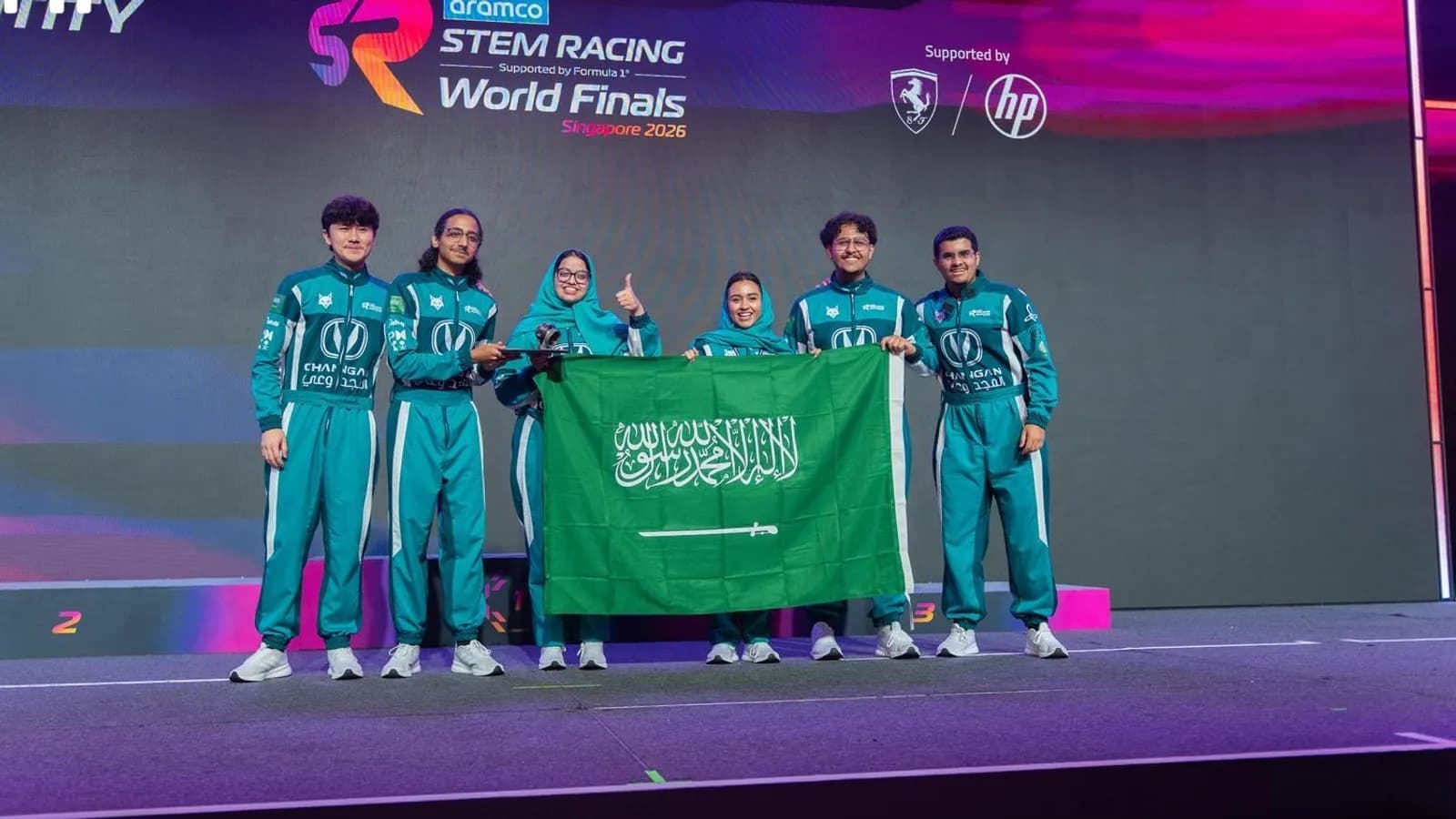 Members of Saudi Team Lynx in their green racing suits holding the flag of Saudi Arabia on the stage of the Aramco STEM Racing World Finals in Singapore, October 2026, a photograph supplied by Ithra