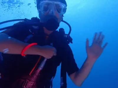 A scuba diver underwater in blue water, hand raised toward the camera, in a 2024 photograph