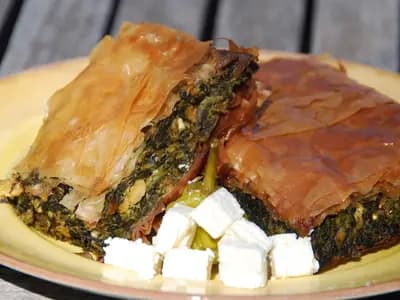 Two wedges of spanakopita on a plate, the layered filo crisp and golden over a dark green spinach filling, with cubes of feta beside them