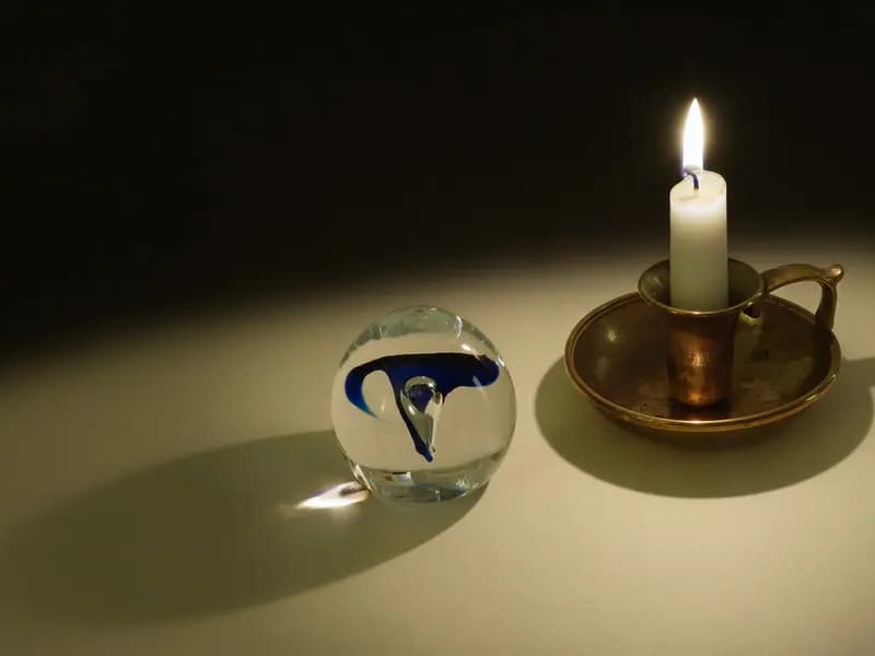 A lit candle in a brass holder on a dark table, its flame refracted through a glass orb beside it