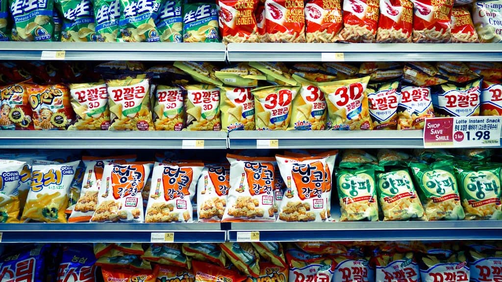 A supermarket aisle shelf stocked with brightly packaged snack products next to a produce section