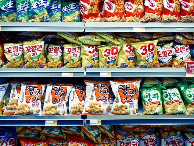A supermarket aisle shelf stocked with brightly packaged snack products next to a produce section