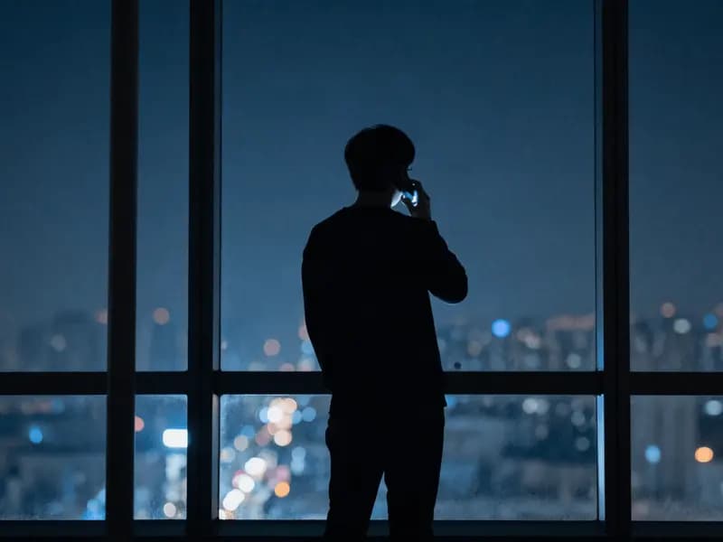 A person on a phone call at a window at night, city lights behind them