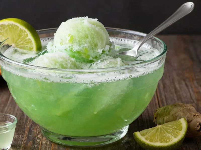 Editorial illustration of a witches brew Halloween punch: a glass bowl of green lime sherbet punch with foamy sherbet scoops, lime slices and a ladle on a dark table.